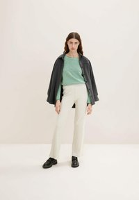 Graue, geknöpfte Jacke über einem strukturierten grünen Oberteil, kombiniert mit cremefarbenen Rippenhosen und schwarzen Schuhen. Minimalistisches Design und neutrales Farbschema.