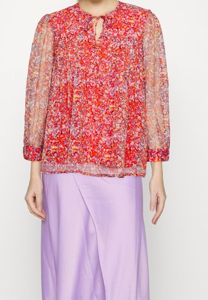 Blouse rouge à fleurs avec des manches transparentes, texture plissée, décolleté en forme de trou de serrure et poignets élastiques, associée à une jupe lavande pastel.