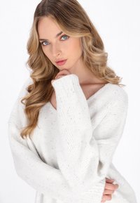 Maglione bianco a maglia con scollo a V, maniche larghe e un motivo testurizzato. Tessuto morbido dall'aspetto accogliente. La modella posa con una mano sul mento.