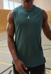 Atleta masculino vistiendo una camiseta deportiva sin mangas de color verde oscuro con el logo blanco de Jumpman, de pie en una cancha de baloncesto cubierta.