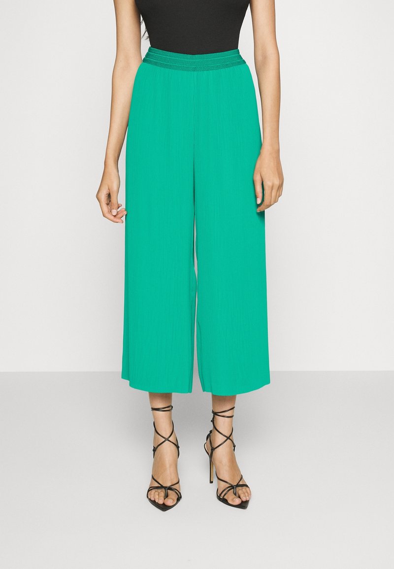 Femme portant un pantalon vert taille haute à jambes larges, associé à des sandales noires à talons hauts et à lanières, debout devant un fond uni.