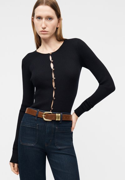 Γυναίκα που φοράει μαύρη ριγωτή ζακέτα με κουμπιά και ανοίγματα σε σχήμα κλειδαρότρυπας και σκούρο μπλε τζιν με καφέ ζώνη, πόζαροντας με το ένα χέρι στη μέση.