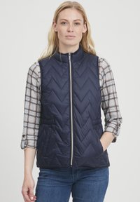Gilet trapuntato blu navy con motivo a zigzag, chiusura con zip e tasche laterali. Indossato sopra una camicia a quadri grigia con maniche arrotolate.