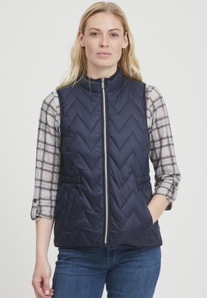 Navy gewatteerd vest met chevronpatroon, ritssluiting en zijzakken. Draagt over een grijsgestreept overhemd met opgerolde mouwen.