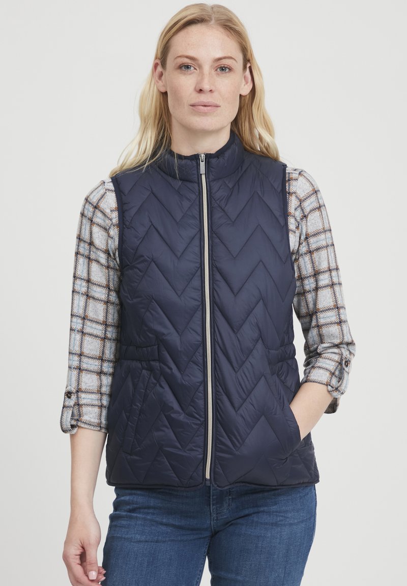 Gilet trapuntato blu navy con motivo a zigzag, chiusura con zip e tasche laterali. Indossato sopra una camicia a quadri grigia con maniche arrotolate.