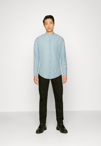 Lichtblauw gestreept overhemd met een mandarin kraag, gecombineerd met zwarte broek en zwarte veterschoenen, staand tegen een effen achtergrond.