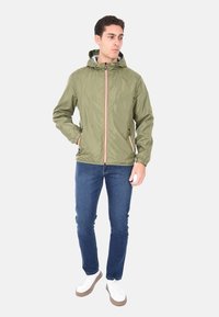 Giacca leggera in nylon verde con cappuccio, chiusura zip frontale e zip a contrasto. Abbinata a jeans blu scuro e scarpe da ginnastica bianche.