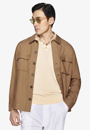 Mężczyzna w beżowej kurtce na guziki, pod którą ma kremową koszulkę polo, białe spodnie oraz przezroczyste brązowe okulary przeciwsłoneczne, patrzący w lewo z jedną ręką w kieszeni.