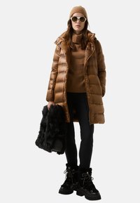 Frau trägt braune gesteppte Jacke, braune Strickmütze, Pullover, schwarze Hose, Stiefel, weiße Sonnenbrille, hält eine schwarze pelzige Tasche, steht vor weißem Hintergrund.