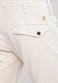 Pantaloni beige in tessuto di cotone con una tasca posteriore con chiusura a bottone e un'etichetta che recita "GUESS 1981."