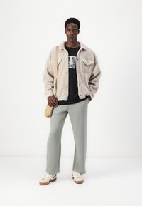 Beige fleece jack, zwart graphic t-shirt, lichtgroene broek, beige schoudertas en witte sneakers met bruine zolen. Simpel ontwerp.