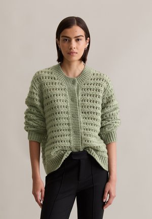 Hellgrüne Strickkarte mit strukturiertem Muster, Knopfverschluss, geripptem Saum und hochgekrempelten Ärmeln, kombiniert mit schwarzen Hosen.