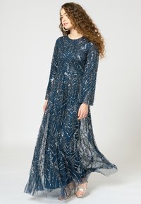 Langes, marineblaues Kleid mit Paillettenblättern, langen Ärmeln und einem fließenden, transparenten Überwurf. Kombiniert mit silbernen High-Heels.