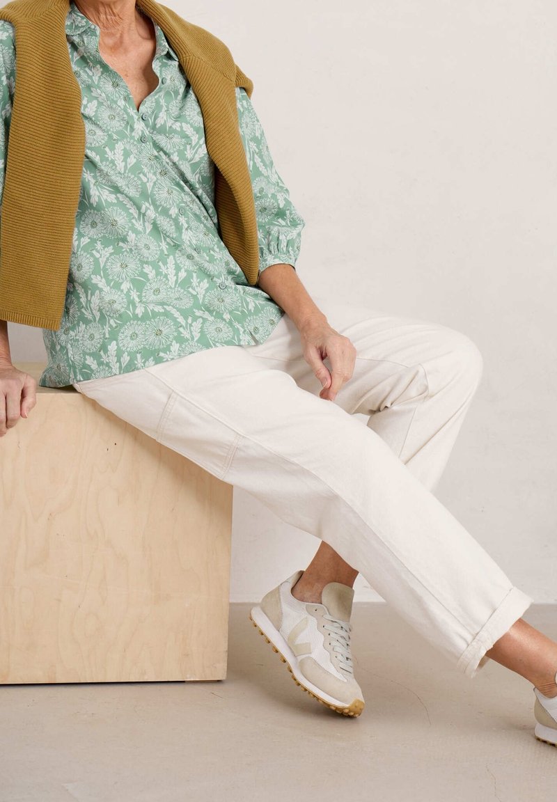 Personne assise sur une caisse en bois, portant une chemise verte à fleurs, un pantalon blanc, des baskets blanches et un pull moutarde drapé sur les épaules.