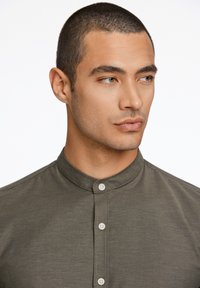 Camisa de manga corta verde oliva con cuello de banda y cierre de botones. Textura suave, detalle de costura sutil y botones blancos.