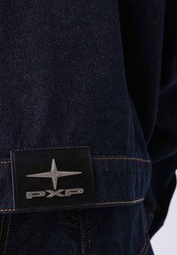 Tessuto di jeans scuro con una consistenza liscia, presenta una toppa in pelle nera. La toppa mostra un logo di stella argentata e "PXP" in caratteri evidenti.