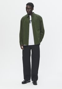 Groene en zwarte gestreepte overhemd, lange mouwen, gedragen over een wit T-shirt, gecombineerd met zwarte broek en schoenen. Eenvoudig ontwerp, casual pasvorm.