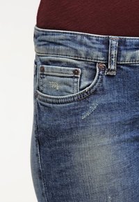 Blå denimjeans som bärs med en burgundy topp, visar detaljerna på framsidan av fickan och bältesöglorna.