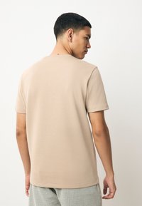 T-shirt beige à manches courtes avec un col rond, texture de tissu lisse et ourlet droit, associé à un pantalon à carreaux, porté de dos.