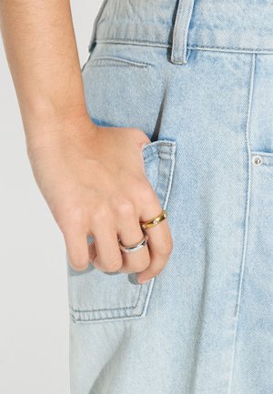 Ręka z dwoma pierścionkami, jednym złotym i jednym srebrnym z wbudowanymi małymi diamentami, spoczywająca na jasnoniebieskiej kieszeni z denimu.