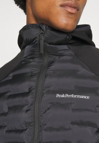 Sort pufferjakke med quiltet tekstur, lynlås og hætte. "PeakPerformance"-logo i hvid på brystet, sat mod et strømlinet design.
