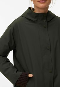 Femme portant un imperméable à capuche vert foncé avec des boutons-pression et une main partiellement dans la poche latérale par-dessus une manche tricotée sombre.