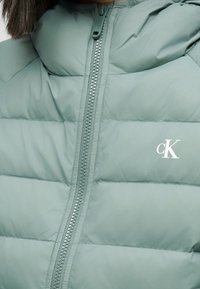 Επένδυση με μαξιλαρωτή υφή σε ανοιχτό πράσινο, με λείο υλικό, διαθέτει φερμουάρ μπροστά και λευκό τυπωμένο λογότυπο στο στήθος.