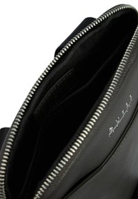 Interior de bolsa de cuero negro con textura suave, cierre de cremallera plateado y forro de tela; presenta un discreto detalle del logo en el interior.