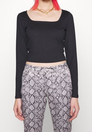 Osoba nosící černý top s dlouhým rukávem a hranatým výstřihem, zlatý řetízek a šedé kalhoty s hadím vzorem.