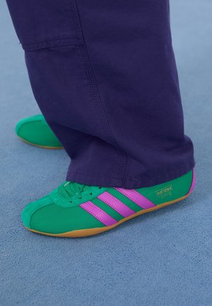 Grønne og pink Adidas Tokyo sneakers båret med mørkelilla bukser på en lysblå tekstureret gulv.