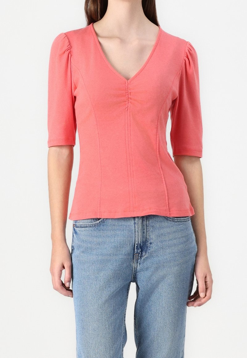 Femme portant un top ajusté rose corail à col en V avec des manches trois-quarts bouffantes et un jean taille haute bleu clair.