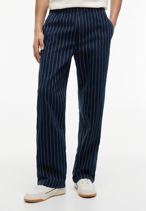 Persona che indossa pantaloni blu navy a righe sottili con le mani nelle tasche e scarpe da ginnastica bianche su sfondo bianco.