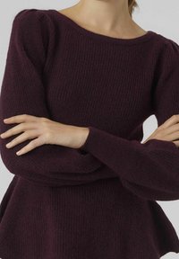 Eine Frau mit verschränkten Armen trägt einen dunkelvioletten gestrickten Langarmpullover mit Rundhalsausschnitt vor einem schlichten, hellen Hintergrund.