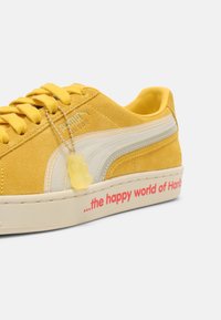 Baskets Puma en daim jaune avec bande blanche, porte-clés ourson en gomme translucide, et texte « the happy world of Haribo » sur la semelle blanche.