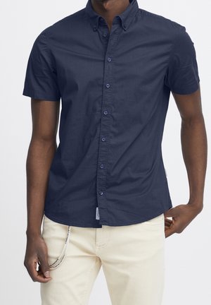 Homme portant une chemise ajustée à manches courtes bleu marine à boutons et un pantalon blanc cassé avec une boucle de cordon de serrage sur le côté gauche.