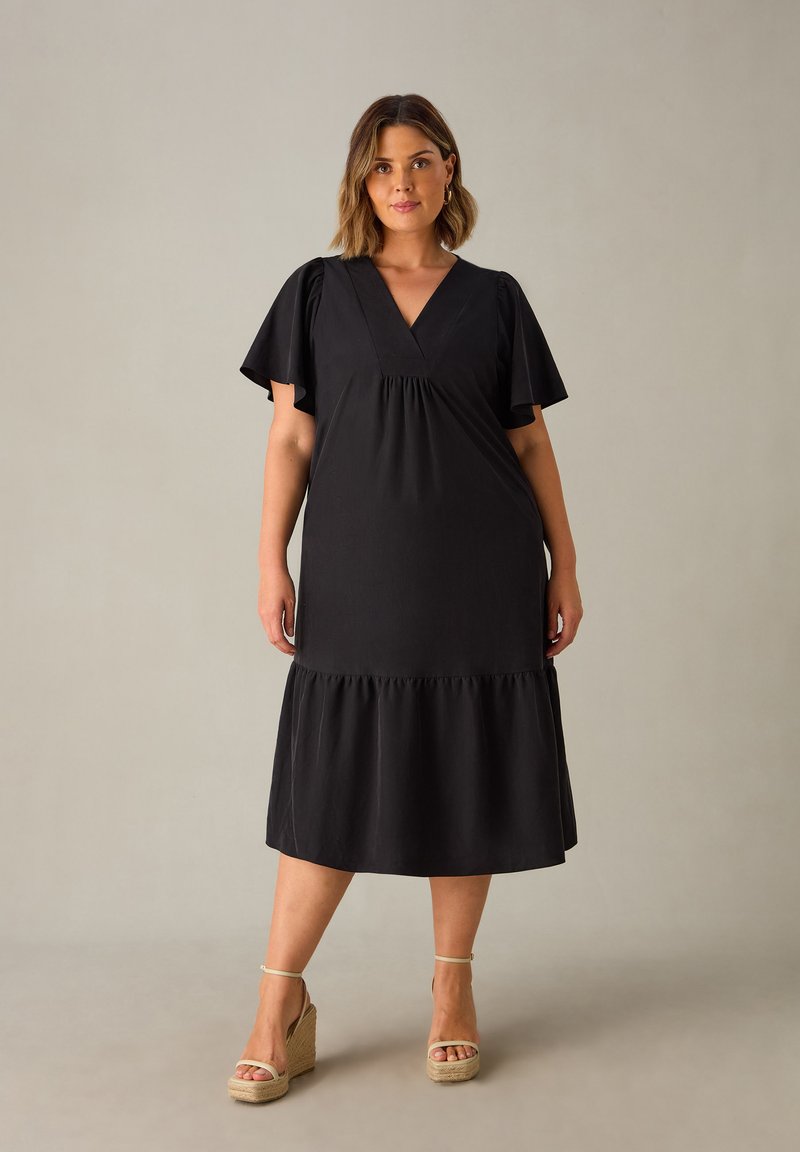 Schwarzes Kleid mit V-Ausschnitt, kurzen, schwingenden Ärmeln und einem gestuften Saum. Das Material ist glatt, mit einer lockeren Passform, die locker fällt. Getragen mit Keilsandaletten.