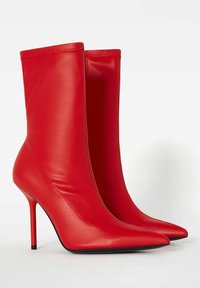 Červené ankle boots z umělé kůže s elegantním, přiléhavým designem, špičatými špičkami a vysokými jehlovými podpatky. Hladká textura, minimální detaily švů.