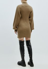 Strickkleid in Braun mit gerippter Textur, hohem Kragen und langen Puffärmeln. Kombiniert mit schwarzen Lederstiefeln über dem Knie.