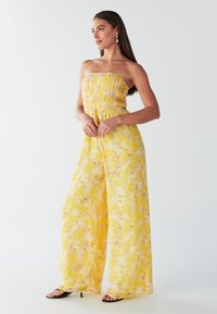 Geltonas gėlių rašto kombinezonas be petnešėlių, laisvo silueto, su plačiomis, laisvai krentančiomis kelnėmis ir surišamu juosmeniu. Lygi, lengva medžiaga.