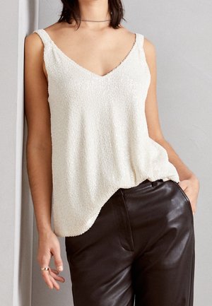 Vrouw draagt een wit, gestructureerd mouwloos V-hals topje dat in een hooggetailleerde zwarte leren broek is gestopt, met een hand in de zak en een zilveren chokerketting.