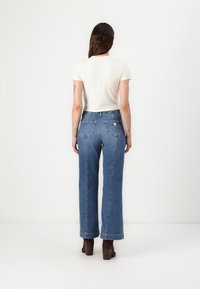 Jeans azules de tiro alto y pierna recta con bolsillos traseros y un acento en forma de triángulo. Combinados con una blusa de manga corta color crema y zapatos de tacón marrones.