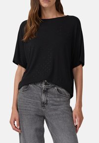 Kobieta ubrana w luźną czarną bluzkę z krótkim rękawem z subtelnymi, małymi ozdobami, włożoną w wysokie, wyblakłe szare dżinsy.