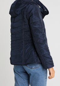 Giacca puffer blu navy con cappuccio, caratterizzata da un design a trapunte, chiusura con zip e un tessuto morbido e lucido. La vista posteriore mostra una silhouette aderente.