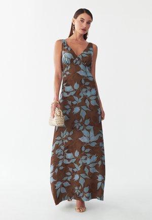 Mujer con un vestido largo marrón sin mangas con patrones de hojas azules, sosteniendo un pequeño bolso tejido y llevando sandalias y joyas doradas.