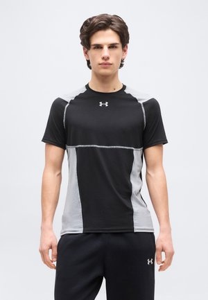 Jeune homme aux cheveux courts bruns portant un t-shirt de sport noir et gris Under Armour et un pantalon noir, debout devant un fond blanc uni.