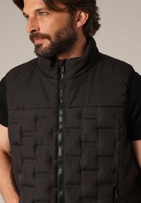 Gilet rembourré noir avec un col montant, un design matelassé à l'avant et un tissu texturé sur les côtés, doté d'une fermeture éclair centrale.