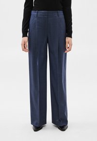 Sieviete valkā plašas tumši zila krāsā drukātas bikses, melnu garām piedurknēm kreklu un melnas ar purngaliem smailas kurpes, stāvot pret vienkāršu fonu.