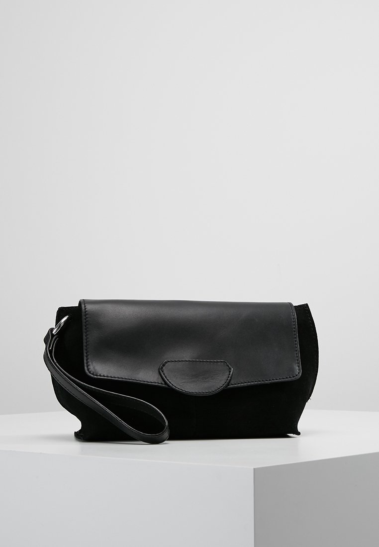 Pochette bracelet en cuir noir avec fermeture à rabat et petite poignée, exposée sur un socle blanc sur fond gris clair.