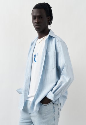 Młody mężczyzna o ciemnej karnacji i dredach, ubrany w jasnoniebieską koszulę w prążki na białą koszulkę z nadrukiem i jasnoniebieskie jeansy, ręce w kieszeniach.