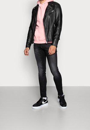 Persoon die een zwart leren jack, een roze hoodie, donkere skinny jeans en zwarte Nike sneakers draagt, staand tegen een vlakke lichte achtergrond.